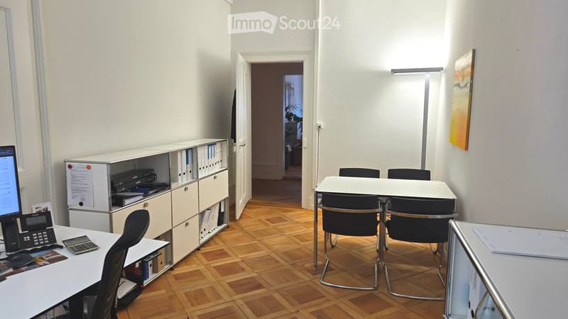Office space with wooden flooring, white walls, and modern furniture including desks, chairs, a screen, telephone, and a cabinet with binders.