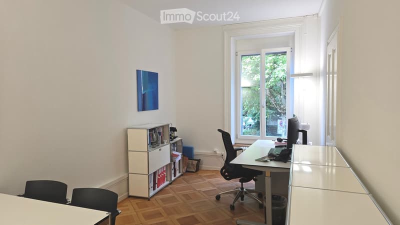 Office space with wooden floor, large window, white walls, and a cabinet with books and files.