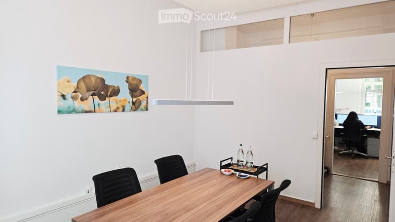 Bright, modern meeting room with a wooden table, black chairs, a large screen, and a picture on the wall. A second room is visible through an open door, containing a desk and computer setup.