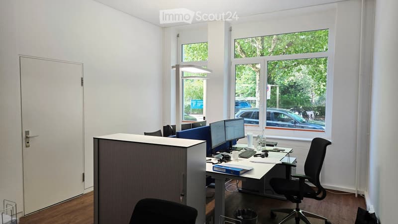 Modern office space with a desk, two computer monitors, ergonomic chair, and filing cabinet. Large windows provide natural light and a view of the street.