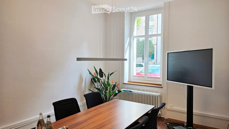 Bright room with a large window, a wooden table surrounded by black chairs, a screen on a stand, a potted plant, and a radiator below the window.