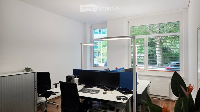 Modern office space with white walls, large windows, and a desk setup with dual monitors, chairs, and office supplies.