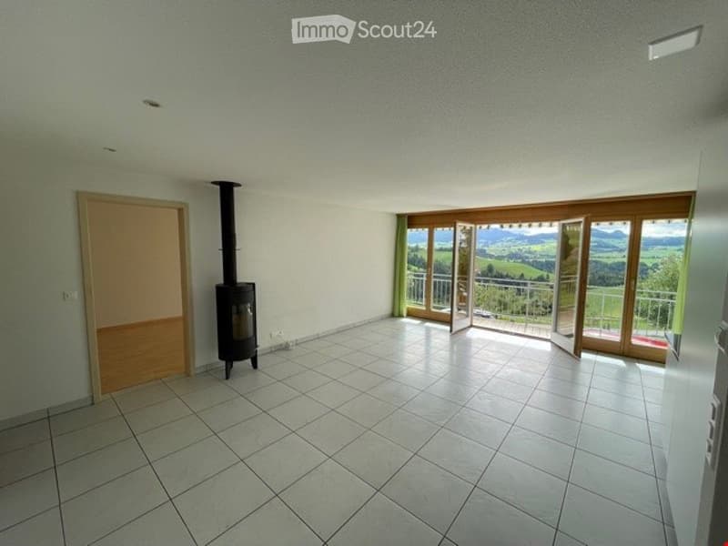 Empty room with tiled floor, white walls, a wood-burning stove, and large windows offering a scenic view of the landscape.