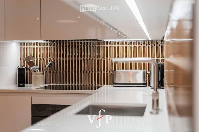 Modern kitchen with light-colored cabinets, a sleek induction stove, a stainless steel toaster, a blender, and a jar on the countertop. The backsplash features a textured, metallic design.