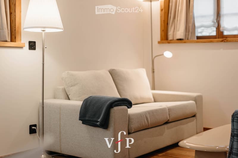 Modern living room with a light grey sofa, a floor lamp, and a small table. The sofa has a dark grey throw on it, and the room features wooden flooring and large windows with wooden frames.
