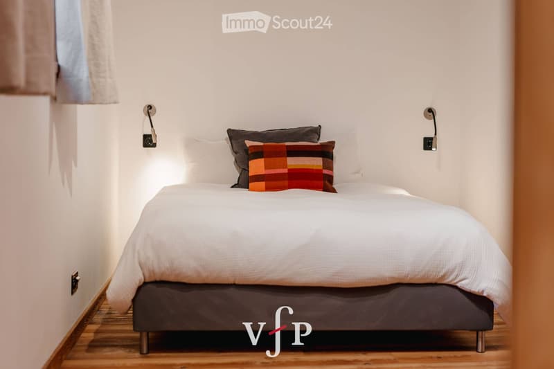 A modern, minimalist bedroom with a neatly made bed, white bedding, and a colorful throw pillow. The room features wooden flooring and wall-mounted lamps on either side of the bed.