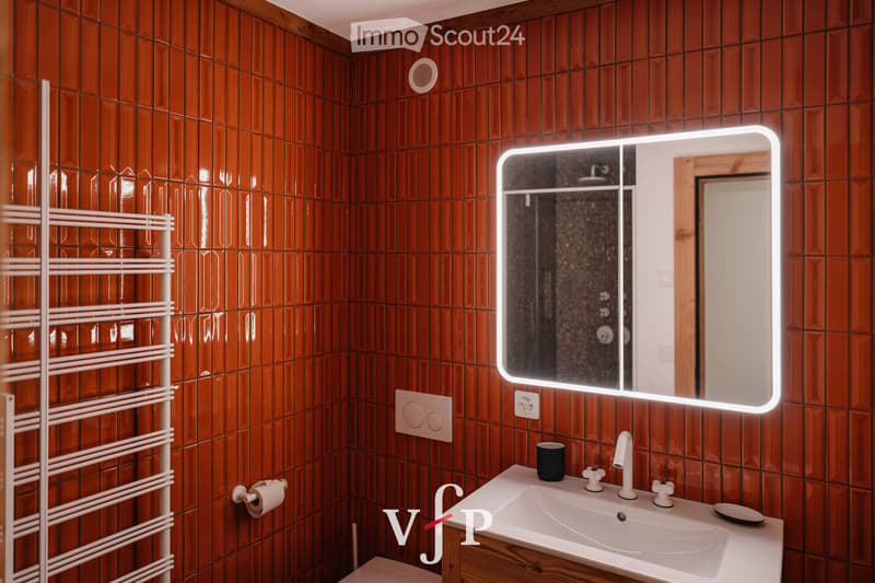 Bathroom with wooden tiled walls, a white sink with a modern faucet, a large illuminated mirror, a towel rack, and a cup on the sink counter.