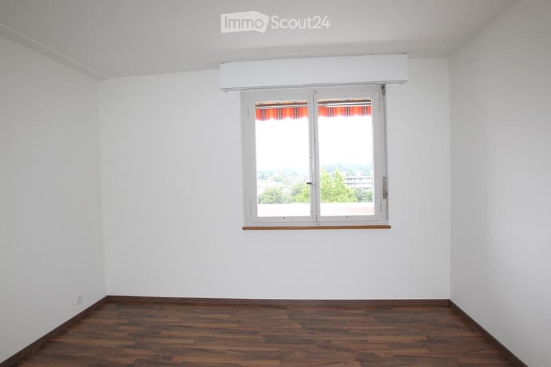 Empty room with white walls, wooden floor, and a large window with a view of the outside.