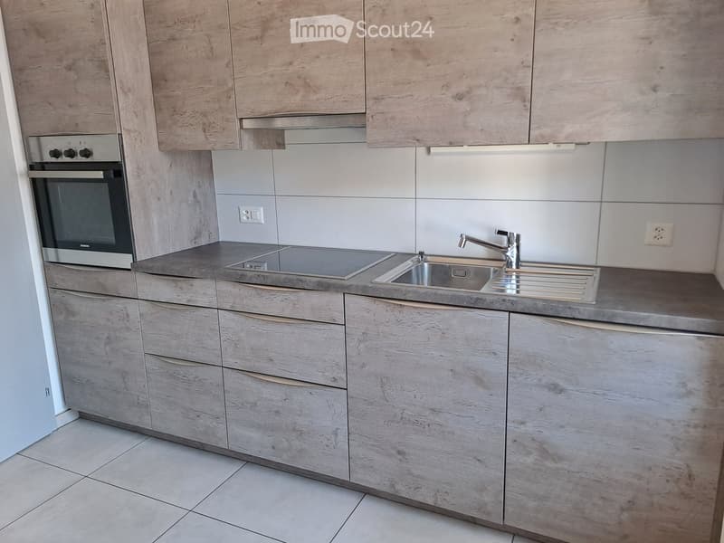 Modern kitchen with light wood cabinets, stainless steel sink, and oven. The countertops are grey, and the backsplash is white tiles.