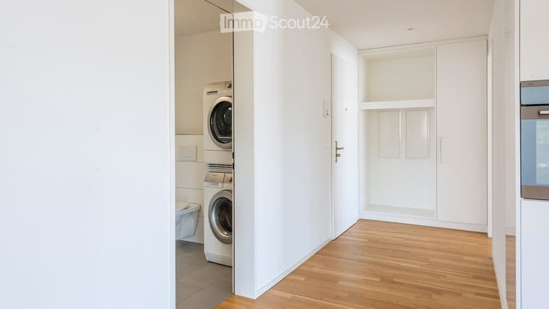 A clean, modern hallway with light wooden flooring, white walls, built-in storage cabinets, and a private washing machine in an adjacent room.