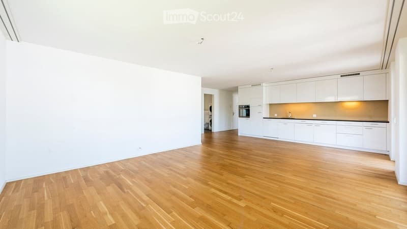 Empty room with light wooden flooring and white walls. Adjacent kitchen area with white cabinets and appliances visible in the background.