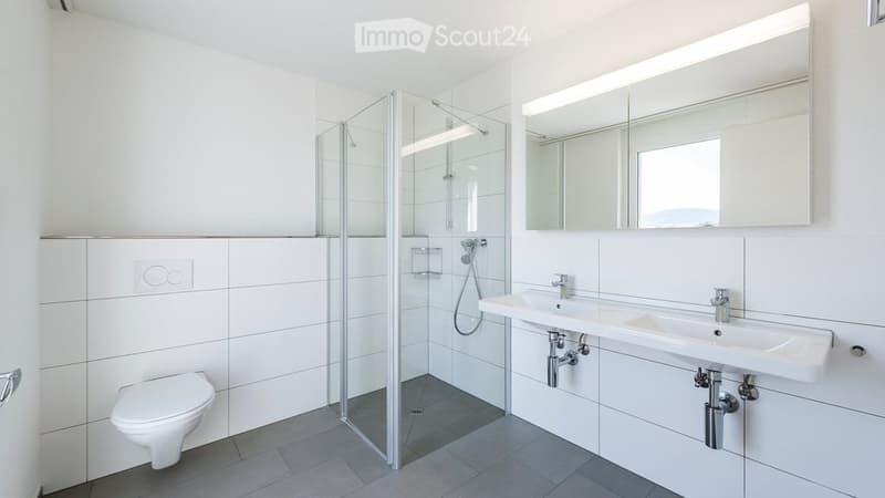 Modern bathroom with white tiles, a glass-enclosed shower, a toilet, a double sink, and a large mirror with built-in lighting.