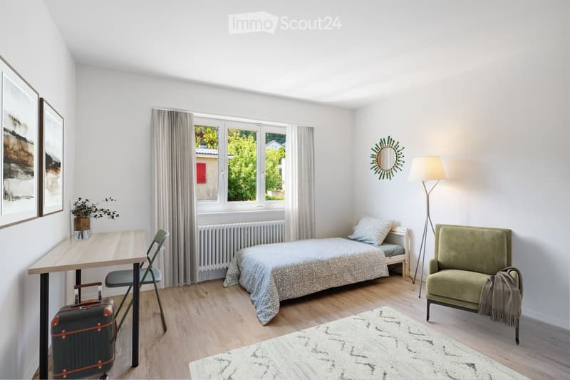A modern, well-lit bedroom with a bed, a desk, a chair, a green armchair, a floor lamp, and a decorative mirror on the wall. The room features wooden flooring, large windows with curtains, and a small plant on the desk.