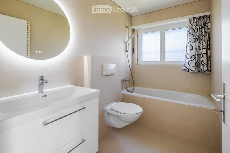 Modern bathroom with a white sink, bathtub, toilet, showerhead, and a large round mirror. The room features light-colored walls and a window with a patterned curtain.