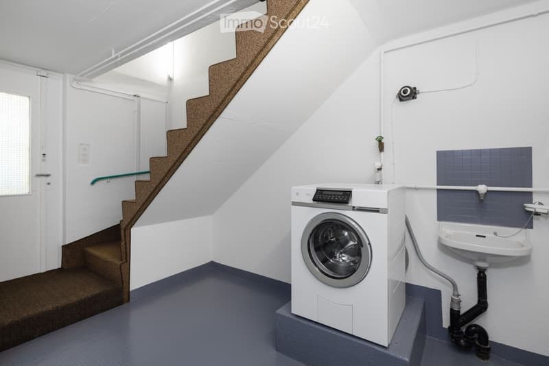 Modern bathroom with a washing machine, sink, and a staircase leading upwards.