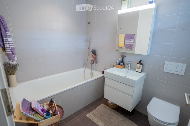 Modern bathroom with a white bathtub, sink, and toilet. Features a built-in mirror cabinet with towels, a small wooden basket with toiletries, and a vase with lavender on the floor.