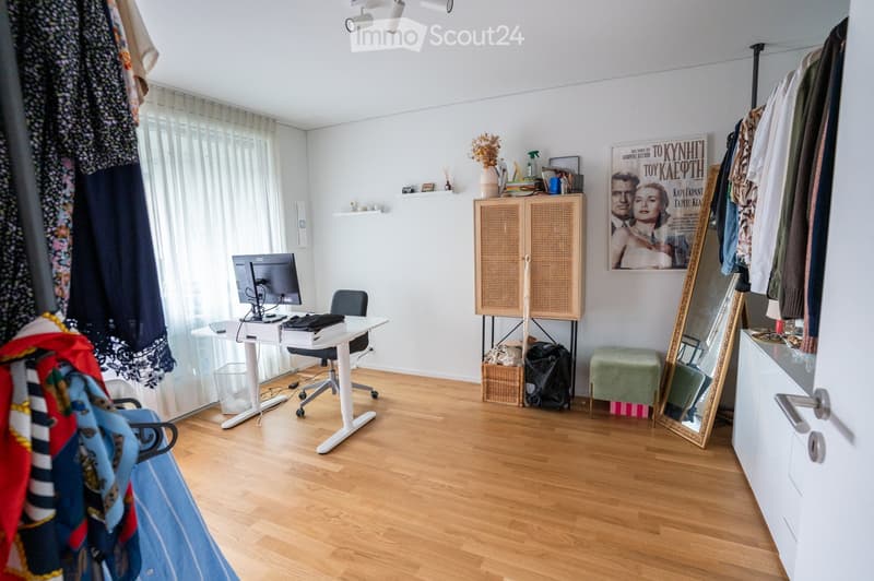 A well-lit room with wooden flooring, a white desk with a computer monitor, a chair, a small cabinet, a green stool, a large mirror, and a clothing rack with various garments.