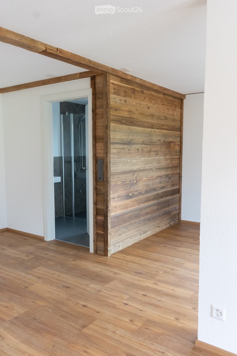 A modern hallway with light wooden flooring, white walls, and a wooden partition leading to a shower area.
