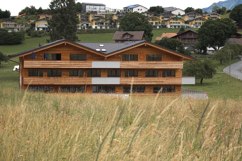 A large, modern wooden building with multiple floors and balconies, surrounded by a grassy field and other residential houses in a hilly landscape.