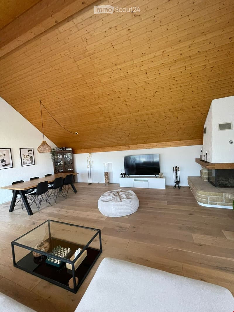 Open-concept living and dining area with exposed wooden beams, hardwood flooring, a fireplace, a large television, a dining table with chairs, a coffee table, and a hanging pendant light.