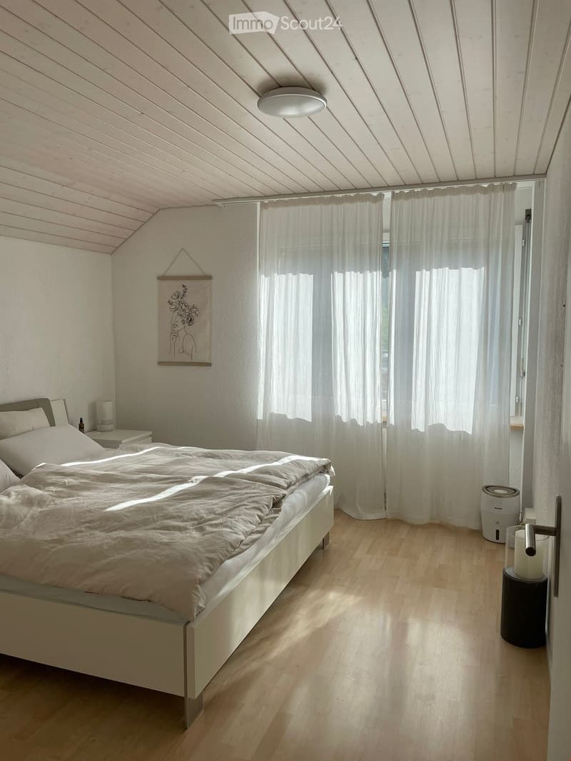 Cozy bedroom with a large bed, nightstand, lamp, and air purifier. Light wooden flooring and white walls with a slanted ceiling.