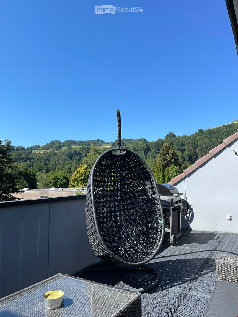 A rooftop terrace with a hanging egg chair, a small table with a candle, and a view of a green, hilly landscape.