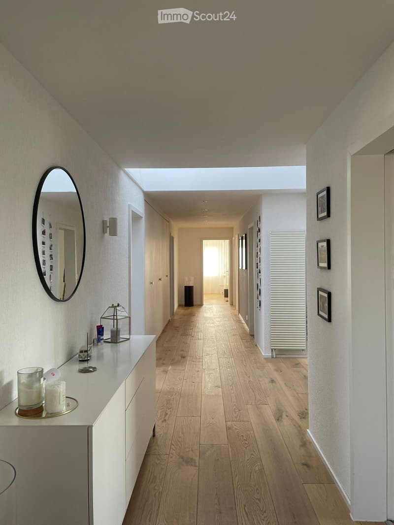 Modern hallway with light wooden flooring, white walls, and a skylight at the far end. Features include a white console table with candles, a decorative lantern, and framed artwork on the walls.