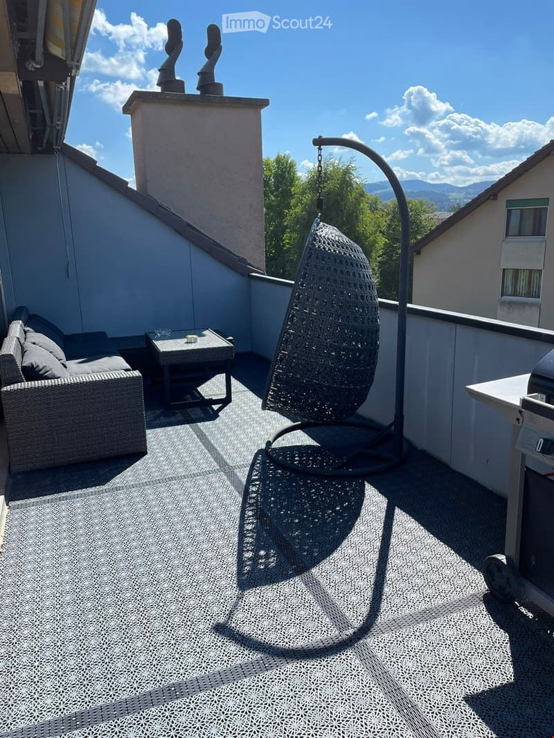 Balcony with a sofa, small table, wicker hanging chair, and grill, overlooking a scenic view with trees and mountains.