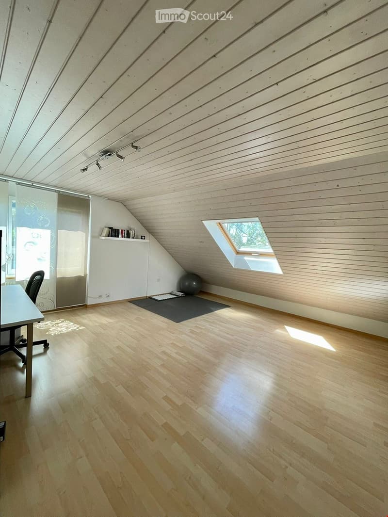 Bright attic room with wooden floors and slanted wooden ceiling, featuring a large skylight window, a small table with a chair, a shelf with books, and a yoga mat with a ball.