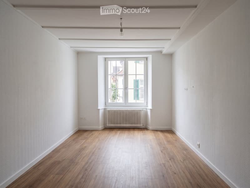 Empty room with wooden flooring, white walls, a window with double doors, and a radiator below the window.