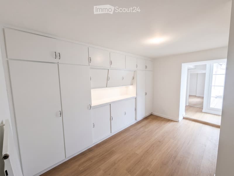 Empty room with white built-in wardrobes and light wooden flooring, featuring large windows and an open doorway leading to another space.