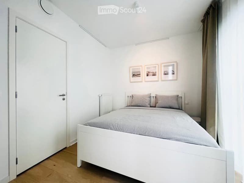 A small, modern bedroom with a white bed, grey bedding, two pillows, and three picture frames on the wall. The room features light wooden flooring and white walls.