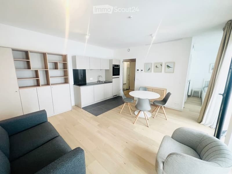 Open-plan living and dining area with a grey sofa, armchair, dining table with chairs, and a compact kitchen featuring white cabinets, a sink, and an oven. Wooden flooring and built-in shelves are visible.