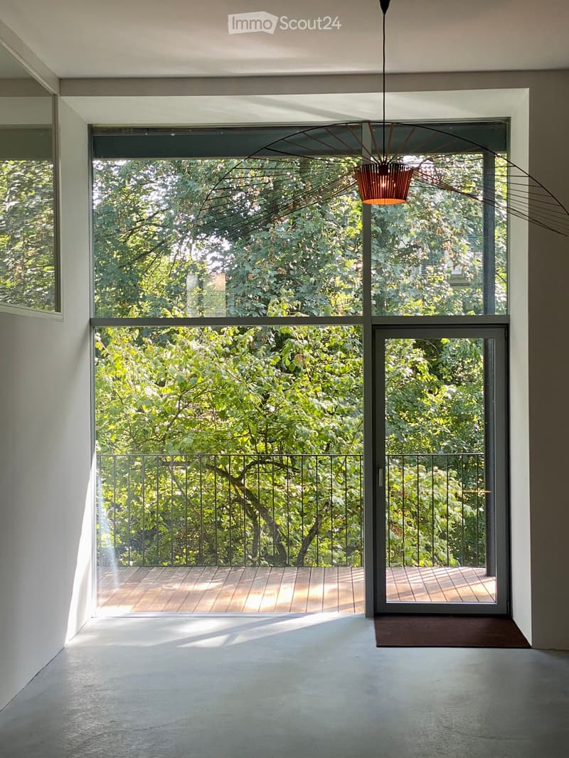 Modern balcony with wooden decking, black metal railing, large glass doors leading to the interior, and a view of lush greenery.