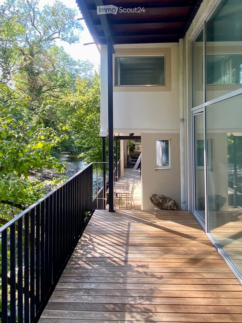 Wooden deck with black railing, overlooking a natural area with trees and a stream. A small table and chairs are placed on the deck.