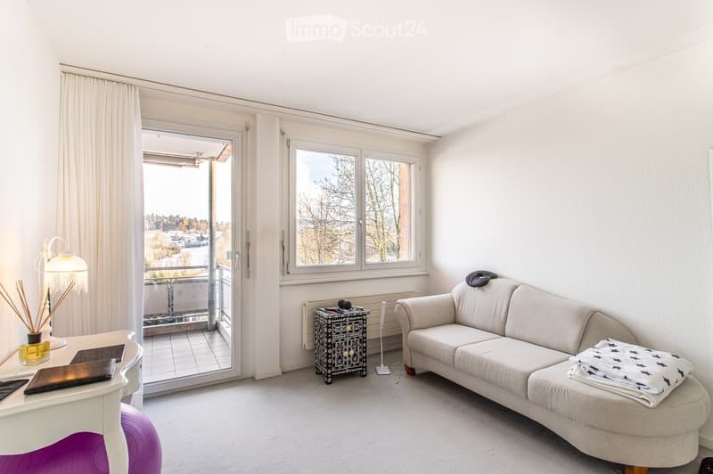 Bright living room with a white sofa, ottoman, small table, lamp, and decorative items. Large windows and a sliding glass door lead to a balcony.