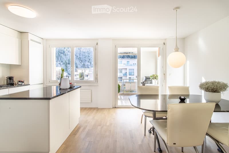 Modern open-plan kitchen and dining area with white cabinets, a black countertop, a glass dining table with four white chairs, and a pendant light hanging above the table. The kitchen features a stove, coffee maker, and a bowl on the counter. Large window