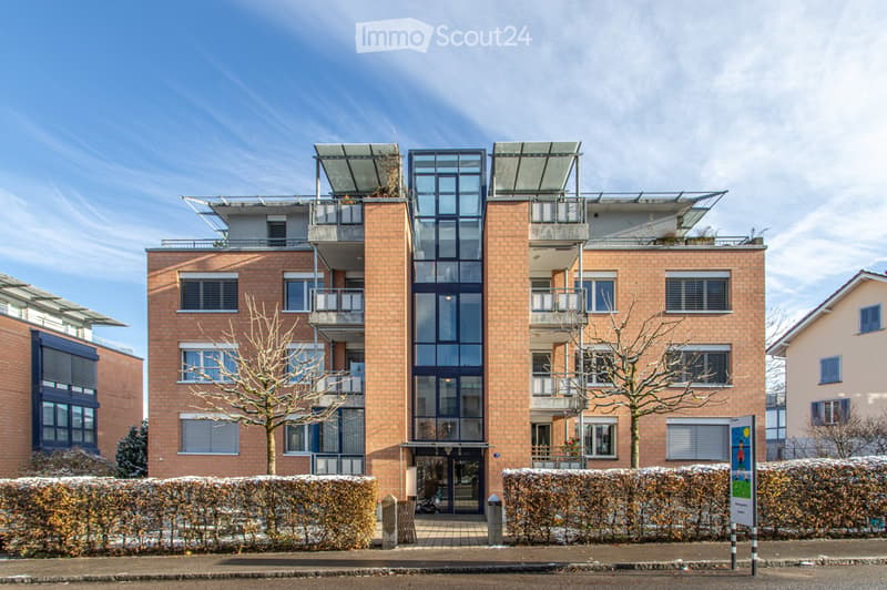 Modern multi-story apartment building with red brick facade, large glass windows, and balconies. The building features a contemporary design with a flat roof and solar panels.