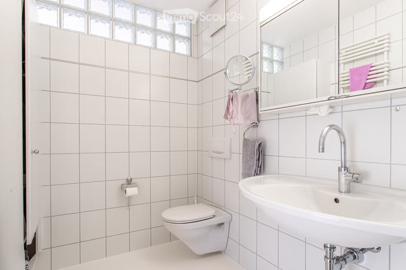 Modern bathroom with white tiled walls and floor, a white sink with a chrome faucet, a toilet, a mirror with a towel rack, and a small window with frosted glass panes.