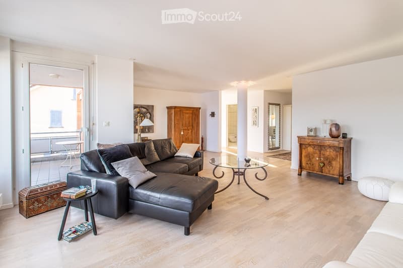 Modern living room with light wooden flooring, a dark leather sectional sofa, glass coffee table, side table with books, and a wooden cabinet.