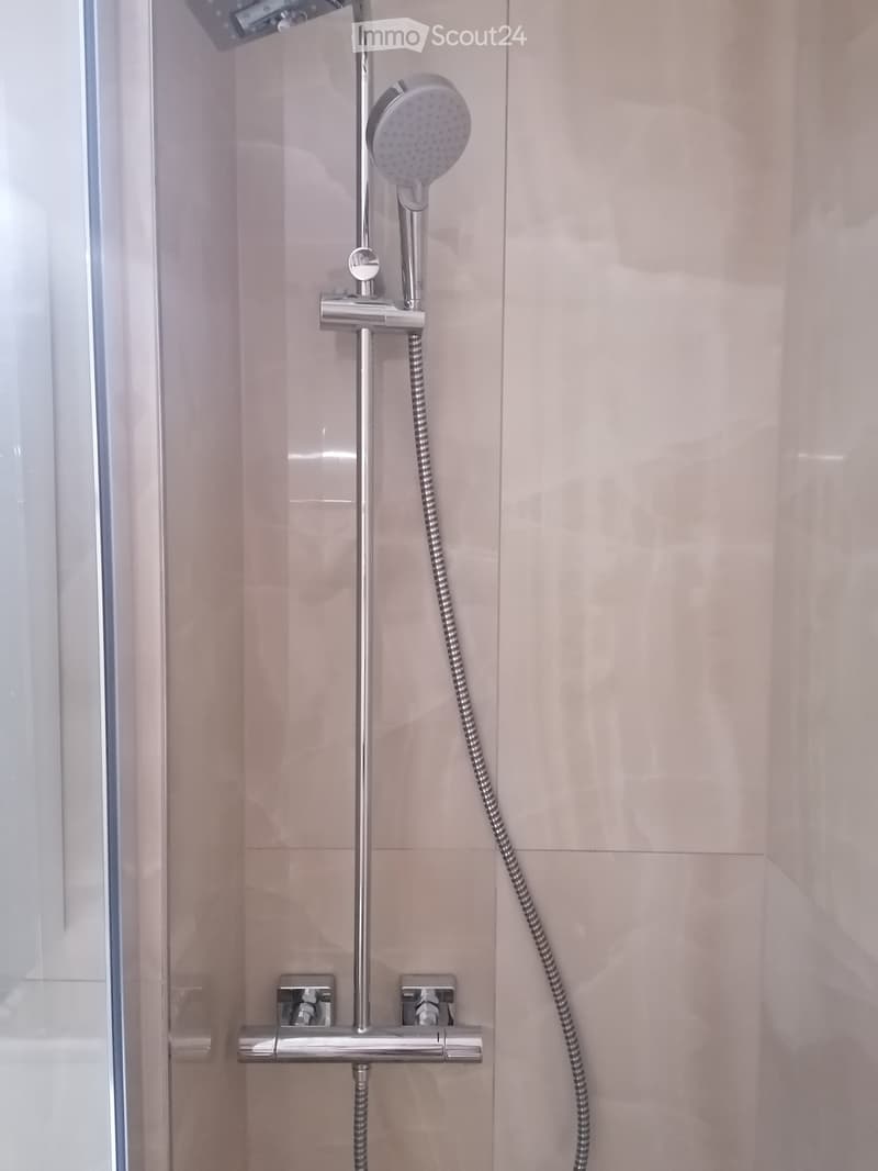 Modern showerhead with handheld hose mounted on light pink tiled wall inside a glass-enclosed shower area.