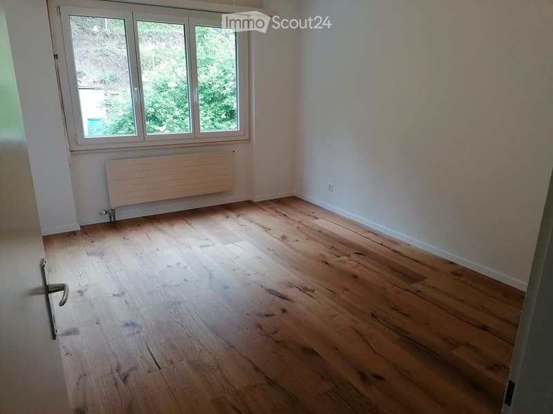 Empty room with light wooden flooring, white walls, and a large window with a view of greenery. A radiator is mounted on the wall below the window.