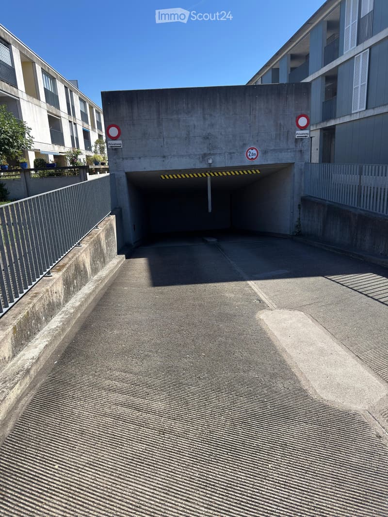 Entrance to a multi-story building with a concrete ramp leading to a covered parking area. The entrance features a sign indicating a private road and a speed limit of 2.0m.