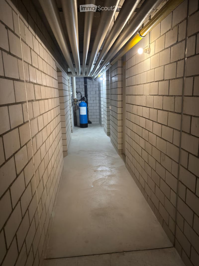 A narrow, industrial-style corridor with brick walls, concrete floor, exposed pipes, and a blue water tank in the background.