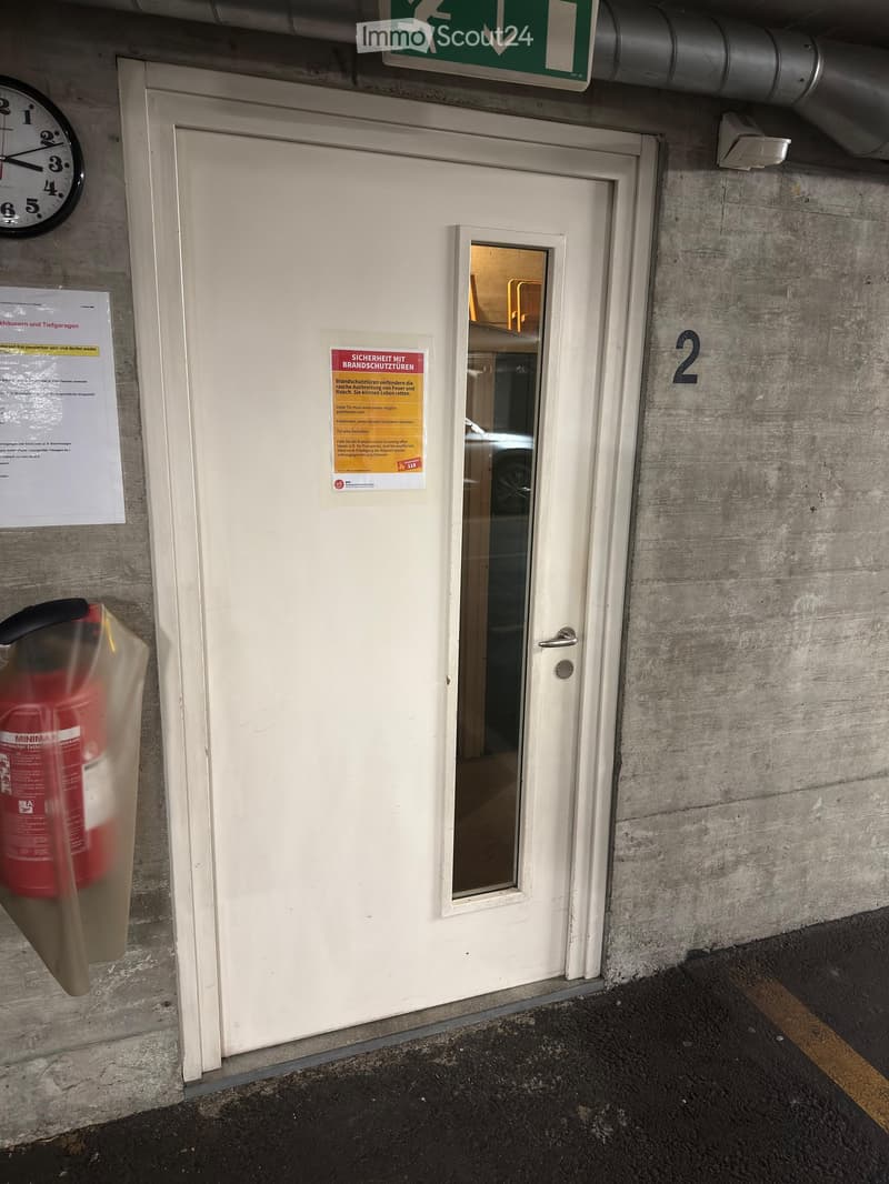 A white door with a small window, a fire extinguisher mounted on the wall, a clock, and a green emergency exit sign above the door.