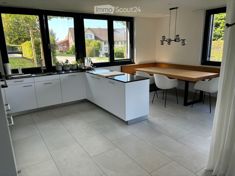 Modern kitchen with white cabinets, a central island, a sink, and a dining area with a wooden table and white chairs. Large windows provide natural light and a view of the garden.