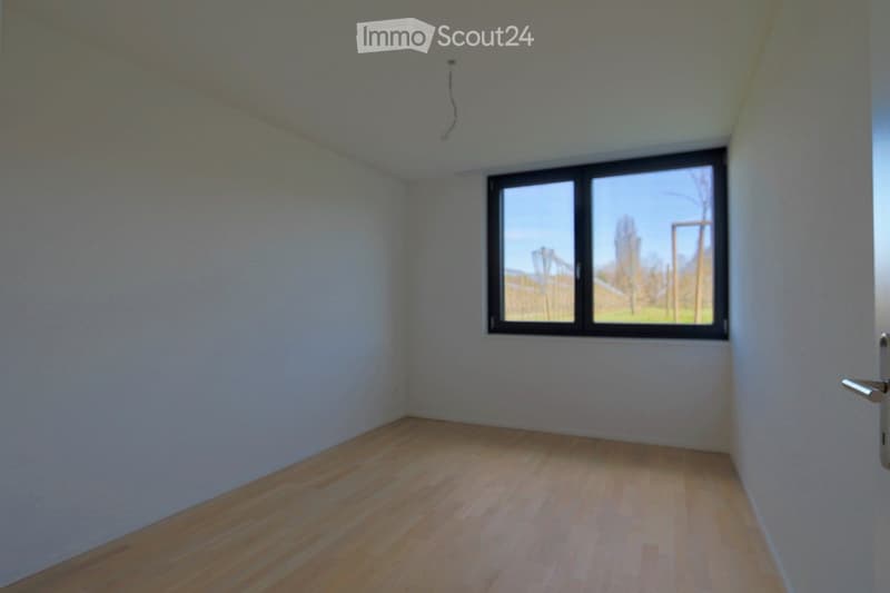 Empty room with light-colored wooden flooring and white walls, featuring a large window with a view of the outdoors.