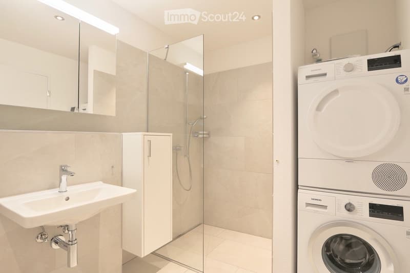Modern bathroom with a sink, mirror, glass-enclosed shower, and stacked Siemens washing machine and dryer.