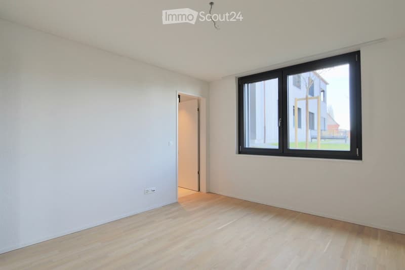 Empty room with light-colored wooden flooring, white walls, and a large window with black frame. There is a doorway leading to another room.
