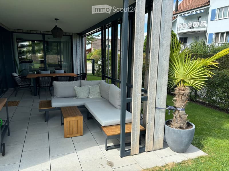 Balcony with a grey sectional sofa, wooden stools, a dining table, and chairs, surrounded by a wooden railing and a potted palm tree.